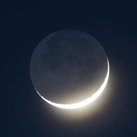 Die überbelichtete zunehmende schmale Mondsichel fast formatfüllend am dunkelblauen Dämmerungshimmel. Die dunkle Seite des Mondes wird als aschfahles Mondlicht sichtbar