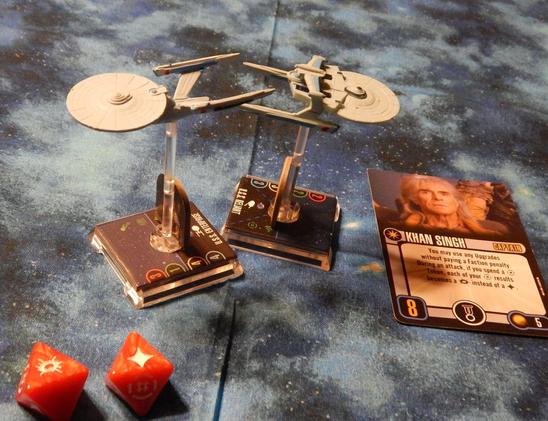 Two game model figures of the USS Enterprise and USS Reliant standing on clear plastic rods out of a base with game stats printed on the cardboard insert. A Khan captain's card sits in front of the reliant with two 8-sided dice (red) showing a hit and critical hit on their faces near the Enterprise. Everything sits on a mottled blue and light-blue nebula starfield cloth.