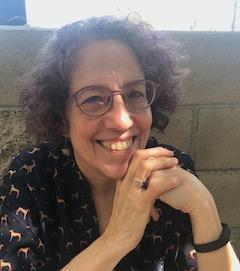 Color photo of author Madeleine E. Robins, a fair-skinned woman with curly brown hair worn in a chin-length bob without bangs. She wears wire-rimmed glasses, a fitness tracker on a black band, a ring on one finger, and a black short-sleeve blouse with a pattern of leggy dogs in profile. She sits in front of a cinderblock wall and rests her elbows on an unseen surface. Strong light comes from over the wall, throwing shadows on her face and making her hair look misty. She looks straight into the camera and smiles. Photo by Rebecca Cavacco.