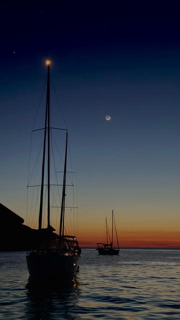 Fast Nacht, die Sonne ist untergegangen. Blick nach Westen, aus einer Bucht heraus in der wir mit dem Boot vor Anker liegen. Links mehrere Segelboote, im Hintergrund die Sichel des Mondes.