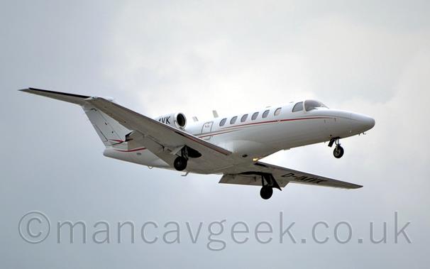 Side view of a white, twin engined BizJet with the engines mounted on the sides of the rear fuselage, flying from left to right at a low altitude, with undercarriage extended and flaps deployed behind the wings, suggesting it is about to land.
The plane is almost entirely white, with a grey and red stripe twisting it's way along the body.
The black letters "VK" can be seen on the engine pod, the rest of the registration obscured by the right wing, although the whole registration, "D-IMVK" can be seen repeated under the left wing.
Flat grey sky fills the rest of the frame.