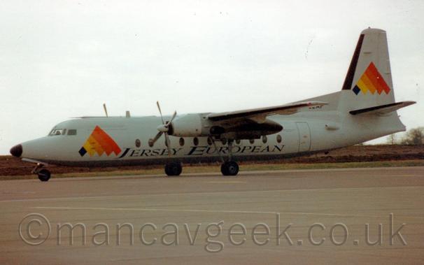 Side view of a white, high-winged, twin propellor-engined airliner taxiing from right to left.
The plane has a white fuselage and brey belly, with a series of black, yellow, orange, and red diagonal stripes arranged to form a triangle on the forward fuselage, a design repeated on the tail.
Black "Jersey European" titles are on the lower fuselage, centred under the wings.
Grey concrete apron fills the foreground, with green grass and brown fields visible under the plane in the distance, under grey sky.