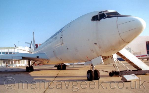 Close-up of the nose of a white, 4 engined jet airliner on display outside a museum, parked facing to the left.
The plane is almost all white, with blue "Air France" titles on the upper forward fuselage.
A small plaque on the ground gives some information about this exhibit, but only a picture of it in flight is discernible here.
A set of white airstairs is connected to the forward door on the opposite side.
A white building stretches across the background, with a pair of military jet trainer aircraft almost appearing to burst out of the roof on the left of the frame. Pale blue sky completes the image.