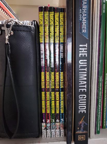 Photo of a colored pencil case and the spines of several books on a shelf; including Transmetropolitan 1-5, DK's The Ultimate Guide to Warhammer 40,000, and several White Dwarf magazines.