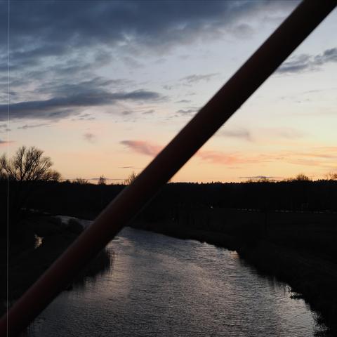 The image shows a scenic view with a river running through the landscape, set against a backdrop of a sunset sky. There are trees silhouetted against the sky, and a large diagonal pole or structure is visible in the foreground. The scene conveys a peaceful and natural atmosphere.