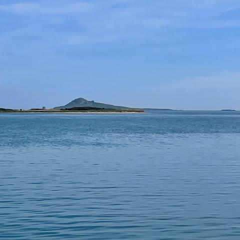 A calm body of water reflects the sky, with a distant island featuring a hill and sparse vegetation. The scene is set under a clear blue sky, conveying a serene atmosphere.