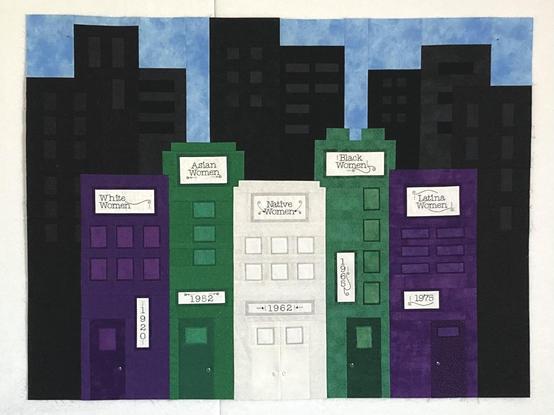 Section of a skyline-themed quilt top, displayed in full on a design wall. The foreground is 5 unique buildings, pieced from left to right in purple, green, grey-ish white, green and purple fabrics respectively, to resemble a Votes for Women flag. The leftmost building has an embroidered sign and address that reads White Women, 1920; the next building has signage that reads Asian Women, 1952. The middle building reads Native Women, 1962, then Black Women, 1965 and the final building is Latina Women, 1975. These addresses signify the year that group of women won the right to vote in the US. In the background are additional buildings, all black on black, set against a light blue sky.
