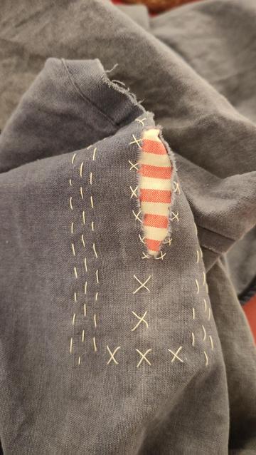 A dark blue pillowcase has a rip along the edge. It is mended: there's a red and white striped piece of an old (and past repair) duvet cover behind the tear which has been attached with white stitching inspired by sashiko. In this case it's mostly rows of running stitches and some crosses.