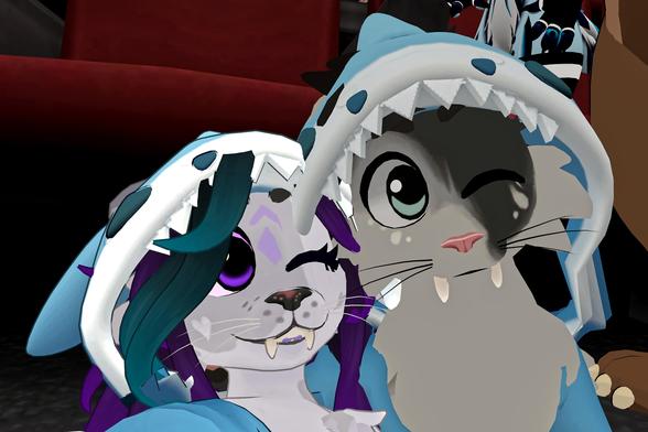 The photo shows two anthropomorphic animal characters, wearing shark-themed hooded outfits, posing closely together. They appear to be in a dark setting, possibly a movie theater. One character is predominantly grey, the other has purple and teal hair. They both seem to be winking at the camera.