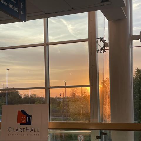 A sunset is visible through large windows, with warm colours illuminating the sky. In the foreground, there is a sign for Clare Hall Shopping Centre. The scene conveys a tranquil atmosphere, with reflections on the glass and greenery outside.