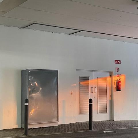 An image of a wall featuring a reflective display case on the left, a double door with slatted vents in the centre, and a fire extinguisher mounted on the right. Two black poles stand in the foreground, with soft orange light reflecting on the wall.