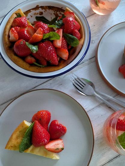 A rectangular photo of a pie, two cocktails, two plates with pie pieces and a fork and spoon sitting on a white wooden background. The pie is a golden-brown colour below a huge pile of bright red strawberries, with green mint leaves poking out between strawberries; there is a light-coloured glaze on top with a few pieces of chopped mint mixed in. It is missing a quarter, which is the slices on the plates - although only one is really shown, as the other disappears out of shot. The cocktails are light-pink coloured sparkling liquids in low-ball glasses, with strawberry quarters and mint leaves mixed in.