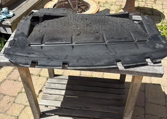 A black plastic under-engine splash panel, removed from my 2003 BMW 5-series station wagon, sits on a weathered wooden work table in my backyard. The leading edge, nearest the camera, has been wiped clean, but most of the panel is coated with a layer of oily, crumbly grime.
