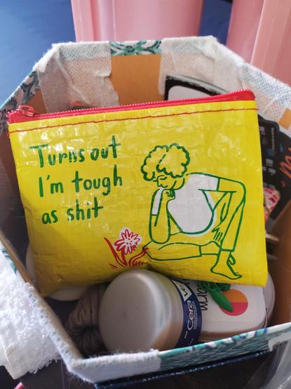A small bright yellow plastic rectangular bag with red zipper, to the right an illustration of a woman sitting cross legged, resting her chin on her hand, gazing at a red flowering plant, and the words "Turns out I'm tough as shit" in upper left quadrant.