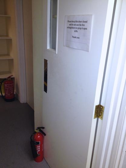 A door, propped open by a fire extinguisher. A paper attached to the door reads: "Please keep this door closed and do not use the fire extinguishers to prop it open with. Thank you"