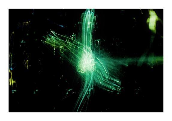 Das Bild zeigt eine abstrakte Lichtmalerei in kräftigem Grün auf schwarzem Hintergrund. Im Zentrum befindet sich ein leuchtender, wirbelnder Knoten aus hellgrünen Lichtspuren. Von diesem Zentrum aus erstrecken sich vier strahlenförmige Lichtbündel in verschiedene Richtungen – oben, unten, links und rechts –, die an Bewegungen von Pinseln oder Laserstrahlen erinnern. Die Lichtlinien sind teilweise geschwungen, teilweise geradlinig, was eine dynamische und fast organische Bewegung suggeriert. Im Hintergrund bleiben vereinzelt schwache Lichtreflexe in Gelb, Blau und Grün sichtbar, was dem Bild Tiefe verleiht.