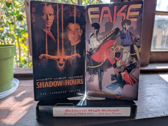 three tapes on a mantel:

SHADOW HOURS
FAKE (an anime)
BELMONT HIGHT SCHOOL CLASSES OF 62/63  35TH REUNION