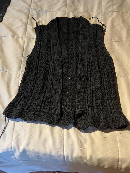 Black lace cardigan sweater body without sleeves yet. It is on a bedspread. There are loose ends. The bottom border is wavy. It has not been blocked.