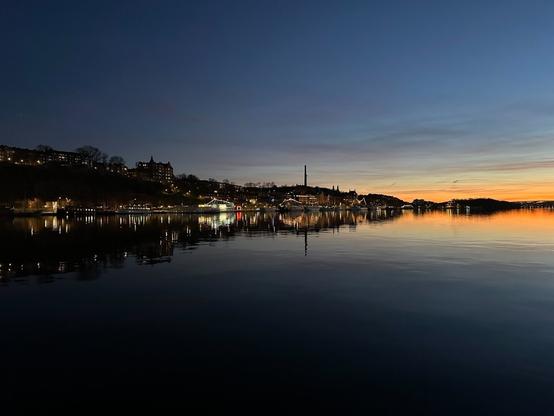 a view over water towards a shore, where boats are moored, and behind them, buildings stand. The shore tapers off to the right of the image, leaving the glow of the setting sun reflecting in the water, and the symmetrical silhouette of a more distant shore.