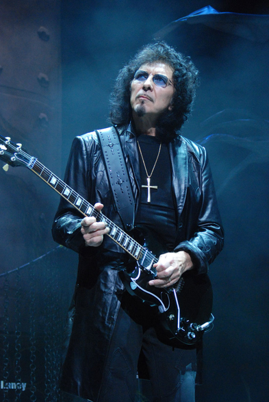 A picture of Black Sabbath guitarist Toni Iommi dressed all in black holding a black Gibson SG guitar and looking off into the distance.