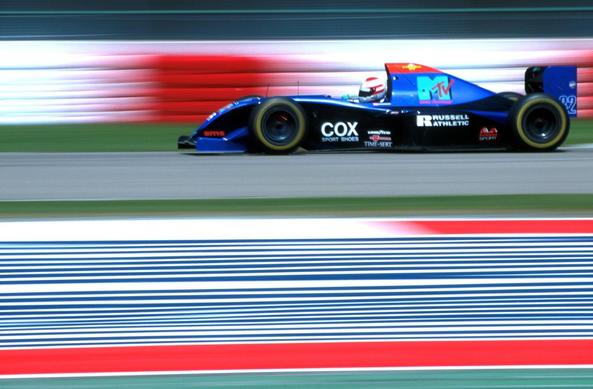 A blue open-wheel Formula 1 car in motion on a racetrack, captured with motion blur. The car features MTV, Cox Sport Shoes, and Russell Athletic sponsorship. The background includes red and white barriers and a striped run-off area with blue, white, and red bands. The driver, Roland Ratzenberger—an Austrian—wears a white helmet with red detailing.