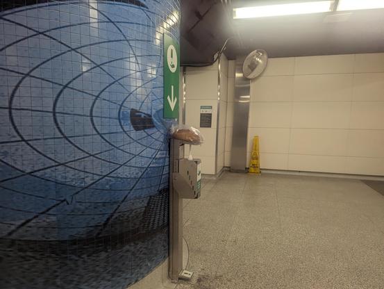 A subway concourse in Toronto.  A hand sanitizer dispenser sits next to a large column covered in a blue and black tile mosaic. A loaf of sourdough bread is left on the hand sanitizer.