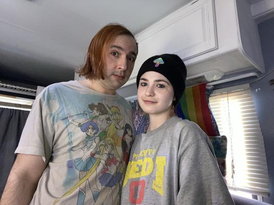 A person with shoulder-length reddish hair wearing a graphic T-shirt featuring anime characters stands next to a person with short hair wearing a black beanie and a gray oversized shirt. They are indoors, with a rainbow flag visible in the background.