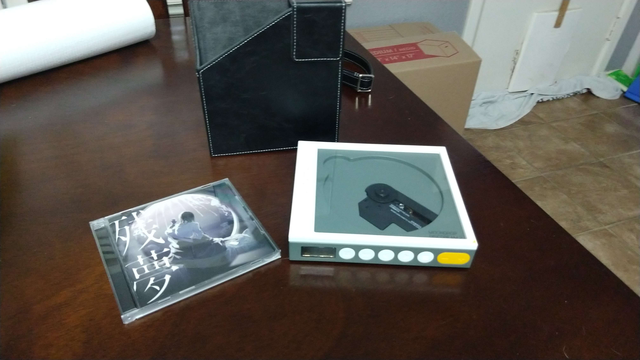 Atop a wooden table is three items laid out: a jewel case with an Ado ablum, the CD player, a Moondrop discdream 2, and a carrying case for it.
