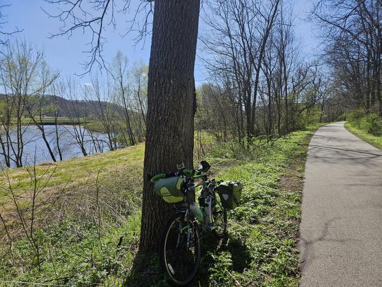A bicycle with green bags leans against a tree. To the left is a blue river, a bluff in the distance is under a blue sky with one small white cloud, to the right is an asphalt trail. The grass is green on both sides. Trees are starting to have some green leaves.