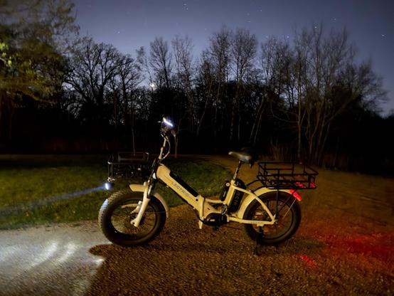 Long exposure photo of a white and black bicycle parked on gravel at night, with no lighting other than the white headlight and red taillight of the bicycle. An area of grass is visible beyond the bicycle, and beyond that a dark tree line. The night sky is visible with the moon at 5% waxing crescent seen through the trees. Some stars are visible in the sky, which is a pale purple due to the long exposure.