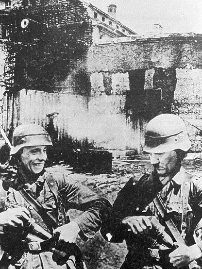 Kaksi taisteluasuista sotilasta, joilla on Joseph Goebbelsin ja Adolf Hitlerin kasvot, seisoo raunioiden keskellä ja asettelee kranaatteja vöihinsä.