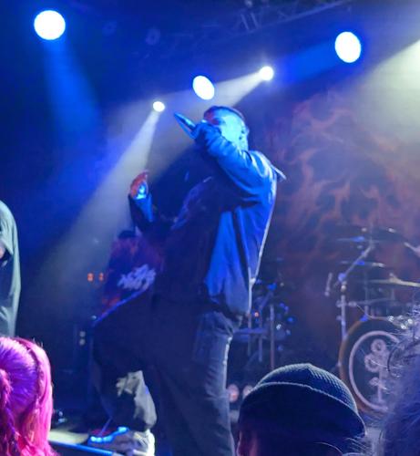 Nasty's lead signer leaning back with his right knee up and microphone at his mouth to capture his vocals, blue lighting around him.