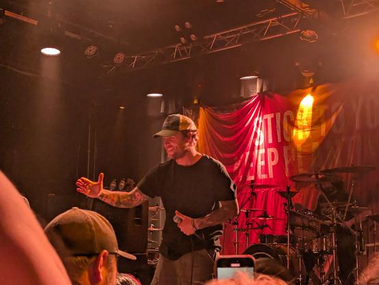 The lead singer for Stick to Your Guns holding out a hand and smiling at the crowd while holding the microphone in his left hand. Red lighting surrounds him.