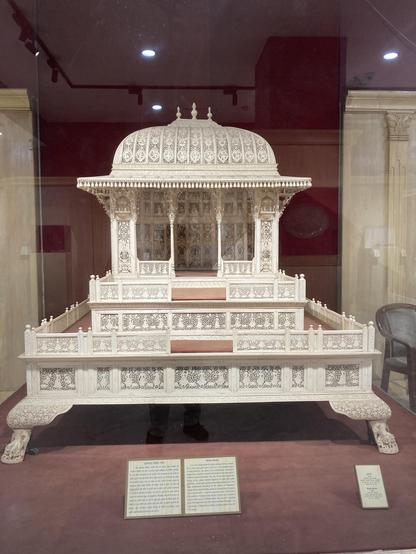 A home shrine, early 20th cent. An intricately carved ivory home shrine having oblong shikhar (top portion of the temple), canopy, front opened garbh ghar (centre portion for keeping idol) placed on two-stepped square wooden platform rests on artistically carved paw-shaped legs. To make smooth movement of this huge shrine, small wheels have been placed intelligently in the inner portion of the legs. A row of tiny bells around abacus of the canopy, bracket figurines on sides and small jharokhas (balcony) on the upper portions of the shrine reminds the Rajasthan palaces' architecture. The size and its grandeur look give the impression that probably this home shrine belongs to the royal family of Rajasthan or Delhi belong to 18th-19th century AD. Simhasana, made of wood is having chhatri, two steps, and four legs. Entire Simhasana is covered with intricately carved ivory plaques, depicting female figures, birds, animals, floral creeper and bells.