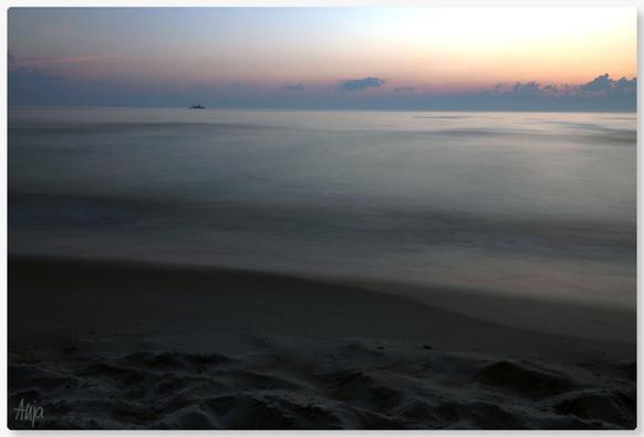 Langzeitaufnahme am Strand der Ostsee. Die Sonne geht unter, Wolken sind zu sehen, ein Schiff fährt in der Ferne in Höhe des Horizonts über das Meer. Die Farben sind gedeckt, von rosa-orange zu blau-grau-braun. Durch die lange Belichtung ist das Wasser verschwommen milchig, der Strand wirkt wie gemalt. 

Long exposure shot on the beach of the Baltic Sea. The sun is setting, clouds can be seen, a ship sails across the sea in the distance at horizon level. The colours are muted, from pink-orange to blue-grey-brown. Due to the long exposure, the water is blurred and milky, the beach looks as if it has been painted.