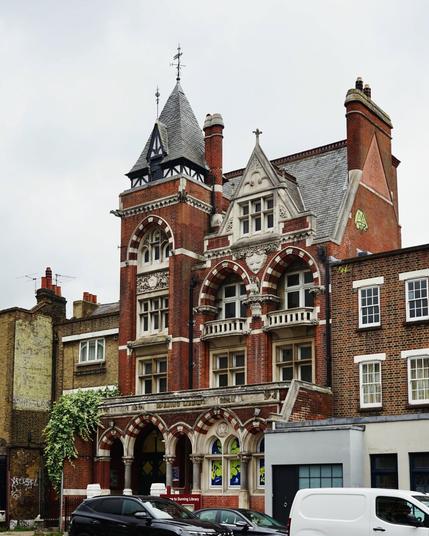 Vue de biais d’un élégant bâtiment en brique rouge de l’époque victorienne. La façade néogothique et asymétrique, sur trois étages, est assez chargée. La partie gauche forme comme une petite tour, mais qui ne dépasse que légèrement de l’ensemble. Il y a des fenêtres à chaque étage, certaines sont surmontées d’un arc en ogive alternant brique et pierre claire. Un faux pignon en pierre vient faire pendant à la tour dans la partie droite de la façade.