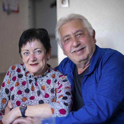 Portrait de Mireille et de son compagnon, ils ont une soixantaine d'année. elle porte un haut gris avec quelques touches de couleurs, il porte un veste qui semble être une veste d'ouvrier, ils sont souriant.