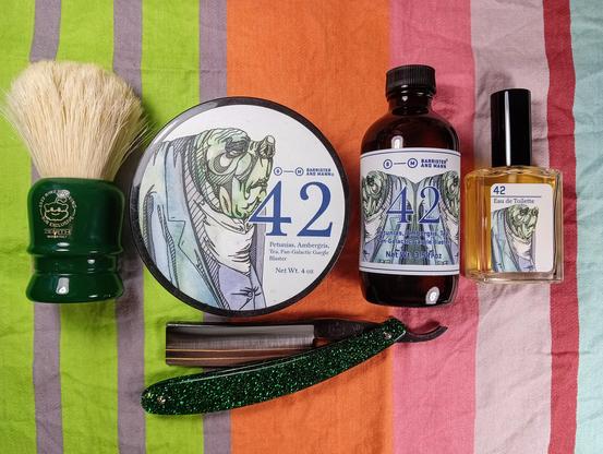 (Le Chat Mistral description)

This image shows a collection of grooming items arranged on a colourful striped cloth. There's a shaving brush with a green handle and white bristles, a round tin labelled "42" with a blue and white design, a brown glass bottle with a similar label, and a small perfume bottle also labelled "42". Additionally, there's a straight razor with a green glittery handle. The items are neatly placed, suggesting a grooming or shaving set.