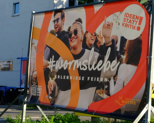Zu sehen ist ein Plakat in der Ortseinfahrt der Stadt Worms, auf der eine Gruppe offenkundig beschwingter Menschen steht, vermutlich tanzend. Bildmittig und im Motiv dominant eine Frau mit einem Weinglas in der Hand. Mittig der Hashtag #wormsliebe, mit dem Claim "Erlebnisse feiern", in der rechten oberen Ecke der Störer "Ideen statt Kritik".