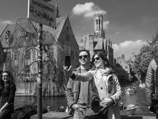 The image is a black and white photograph capturing a moment by a canal in Bruges, Belgium. In the foreground, a couple is taking a selfie. The man is wearing a dark jacket and sunglasses, while the woman is dressed in a light-colored trench coat, a scarf, and sunglasses, holding a smartphone. Behind them, traditional European architecture with steep roofs and a prominent tower is visible, suggesting a picturesque setting. The canal is bustling with boats, and other people are seen enjoying the scenery. The atmosphere is lively, with a clear sky and a few clouds.
