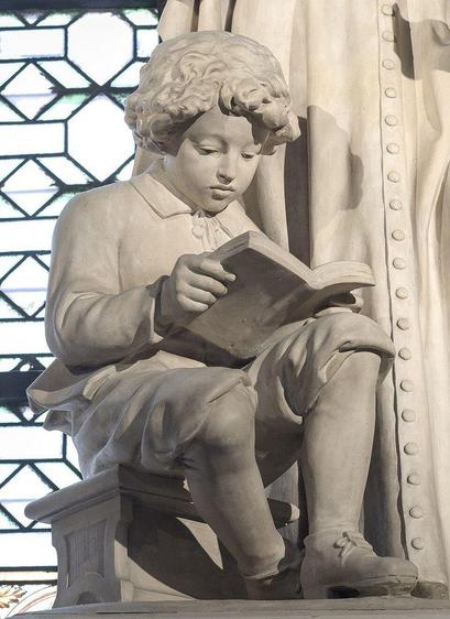 Détail d'une statue de saint Jean-Baptiste de La Salle réalisée par Alexandre Falguière, située dans l'Église Saint-Sulpice de Paris. Cette sculpture représente un enfant assis, vêtu de vêtements d'époque, concentré sur la lecture d'un livre qu'il tient ouvert dans ses mains. Une fenêtre au motif géométrique en arrière-plan complète la scène.