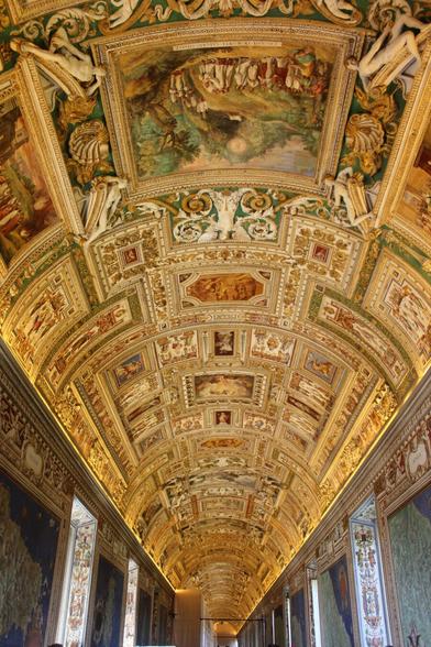 A detailed view looking up at the elaborately decorated and painted barrel-vaulted ceiling of a long hallway, featuring numerous frescoes, sculptures, and ornate architectural details in gold, green, and cream colors.
