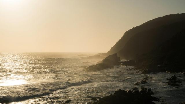 Golden hour sunlight illuminates a rocky coastline with waves crashing against the shore.