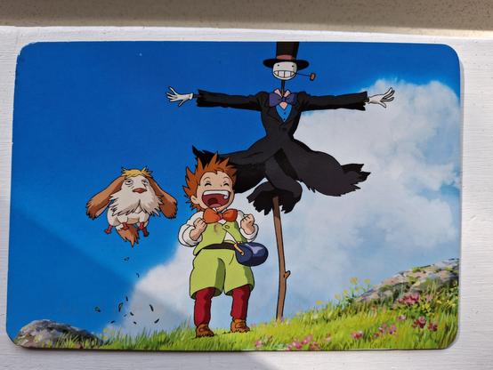A postcard of a still from the animated film Howls Moving Castle. On the side of a grass and flower covered hill are a small, old looking shaggy dog jumping in the air, a small boy dressed in green smiling, and a scarecrow with a pipe and top hat.