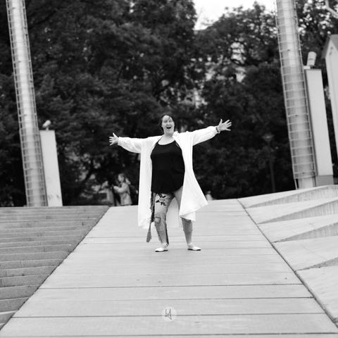 A person standing on a paved walkway with arms outstretched, wearing a long white coat, a black top, and embroidered jeans. The black-and-white image features trees and architectural elements in the background, including tall cylindrical structures and steps.