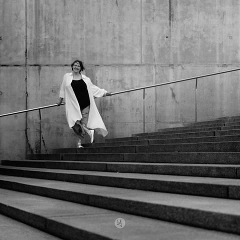 A black and white photograph of a person descending concrete stairs. They wear a long white coat over a black top and ripped jeans. A metal handrail runs alongside them, and the background features a textured concrete wall, emphasizing a minimalist, urban style.