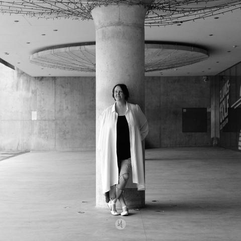 A black and white photograph of a person standing next to a large concrete pillar in an architectural, urban setting. They wear a long light coat over a dark top and jeans, paired with white shoes. To the right, a sign reads 'EL MUSEO DEL GOLF,' framed by the surrounding concrete walls and circular structures in the background, adding texture and visual interest to the composition.
