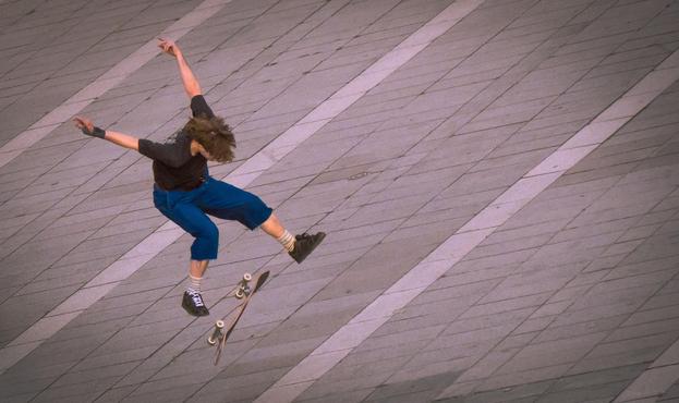 La photographie représente une personne effectuant une figure sur un skateboard. L'individu est en l'air, les bras écartés vers l'arrière, le skateboard basculant en dessous. L'arrière-plan est constitué d'une surface carrelée.
