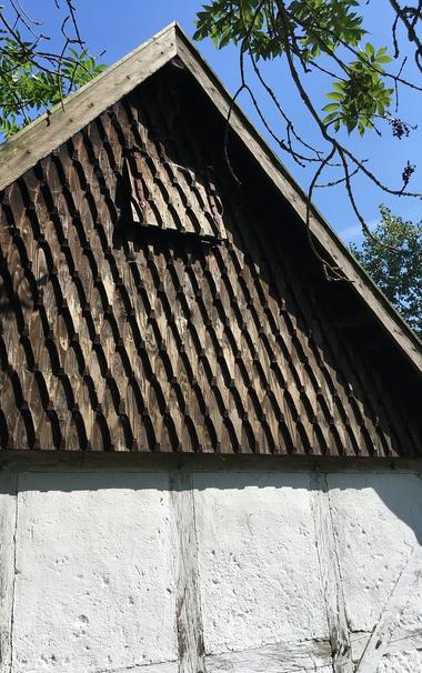 Foto: Ausschnitt eines weiß getünchten kleinen Fachwerkhauses; der Giebel ist vollständig mit recht kleinen, dunkelbraun gebeizten Holzschindeln behängt, die ähnlich einer Pfeilspitze gesägt und gefräst sind. Oben an der Giebelspitze befindet sich ein kleiner Fensterladen, der ebenfalls mit jenen Holzschindeln besetzt wurde.
