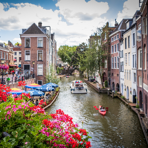A scenic canal in Utrecht, the Netherlands, lined with historic brick buildings, flower-covered terraces, and outdoor cafes. A white tour boat and a red kayak float on the water, while vibrant red and pink flowers bloom in the foreground.